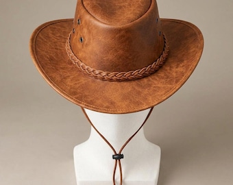May include: A brown hat with a textured finish, featuring a wide brim and a braided band around the crown. The hat includes metal eyelets on the sides for ventilation and a thin brown chin cord with an adjustable black plastic toggle fastener.