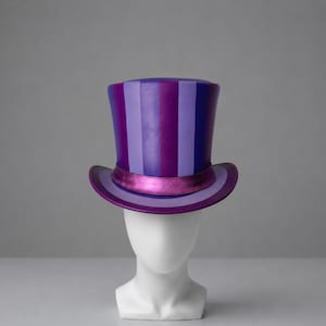 Puede incluir: Un sombrero de copa con rayas verticales en tonos alternos de morado oscuro y lavanda claro. El sombrero incluye una banda de color magenta metálico que rodea la base de la copa y un ala ancha y curva a juego con los tonos morados y lavanda.