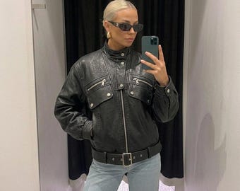 May include: A black leather jacket featuring a high collar, a front zipper closure, and a buckled belt at the hem. The jacket includes two chest pockets with silver-toned zippers and snap buttons, paired with light blue denim jeans.