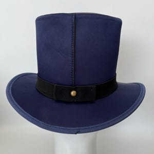 Puede incluir: Un sombrero de copa azul marino con una banda negra y un bot&oacute;n dorado. El sombrero tiene un ala ancha y una corona alta y cil&iacute;ndrica. El sombrero est&aacute; hecho de una tela lisa y tiene una costura cosida en el centro.