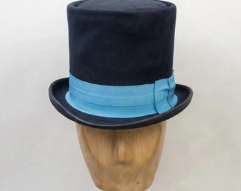 May include: A dark navy blue top hat featuring a flat crown and a wide, curved brim. The base of the crown is encircled by a bright light blue grosgrain ribbon band that is tied in a small, folded bow detail on the side of the hat.