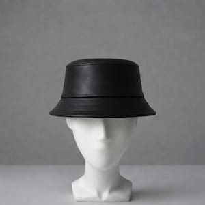 Puede incluir: Un sombrero de pescador negro liso con un acabado mate y suave. El sombrero presenta una copa redondeada y un ala inclinada hacia abajo con tres líneas de costura horizontales que rodean la base de la copa, ofreciendo un aspecto estructurado.