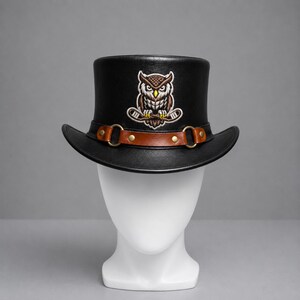 May include: A black top hat featuring an embroidered owl graphic holding two hockey sticks. The hat is accented with a brown leather band secured by two gold-toned metal rings and rivets, creating a structured look with a wide, curved brim.