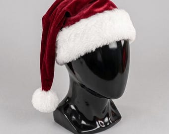 May include: A deep red velvet Santa hat featuring a thick, white faux fur trim around the base and a matching white faux fur pom-pom at the tip. The hat is displayed on a smooth, black, featureless mannequin head against a neutral background.