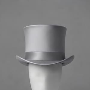 Puede incluir: Un sombrero de copa de color gris claro sólido con un acabado mate suave y una banda de cinta de satén gris a juego que rodea la base de la copa. El sombrero tiene una copa cilíndrica estructurada y un ala curva que se eleva a los lados.