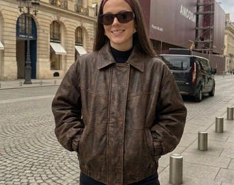 May include: A brown leather bomber jacket with a collared neckline, long sleeves, and a front button placket. The jacket features a distressed texture, ribbed cuffs, and a ribbed hem. It is worn over a black mock neck top with dark sunglasses.