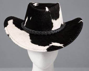 May include: A cowboy hat featuring a black and white cow print pattern with a textured hair-on-hide finish. The hat includes a black braided band encircling the base of the crown and a wide, curved brim that slopes downward on the sides.