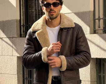 May include: A dark brown leather jacket with a thick cream-colored shearling lining, a wide fold-over collar, long sleeves with shearling cuffs, a front zipper closure, and adjustable buckle straps at the collar and waist, worn over a white t-shirt.