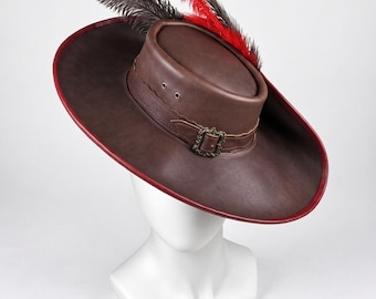 May include: A wide-brimmed brown leather hat featuring a red and dark gray feather plume. The hat includes a decorative metal buckle on a leather band, stitched detailing, and two silver-toned metal eyelets on the side of the crown.
