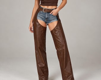 May include: Dark brown leather chaps featuring a wide waistband with a silver-toned metal buckle closure. The chaps are worn over blue denim shorts, extending down the legs in a straight-cut design with a smooth, matte finish.