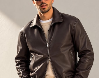 May include: A dark brown leather jacket with a classic collar, a full-length silver metal zipper, and ribbed cuffs and hem. The jacket is worn over a beige ribbed sweater and is paired with dark gray trousers and black rectangular sunglasses.