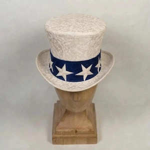 Puede incluir: Un sombrero de copa con corona y ala de encaje blanco. Una banda ancha de color azul oscuro rodea la base de la corona, decorada con un patrón repetitivo de grandes estrellas blancas de cinco puntas. El sombrero se exhibe sobre un soporte de madera.