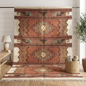 Op de afbeelding: Een groot rechthoekig vloerkleed met een herhalend geometrisch patroon in tinten terracotta, roest, saliegroen en crème. Het kleed heeft een verweerde textuur met horizontale banden en een gefranjerde rand aan de onderkant.