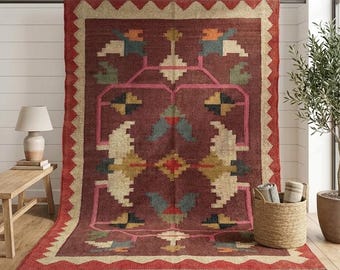 Könnte beinhalten: Ein großer rechteckiger Teppich mit tiefrotem Hintergrund und einem geometrischen Muster in Creme, Blau, Orange und Gelb. Der Teppich hat einen breiten roten Rand mit Zickzack-Kante und Fransen am unteren Ende sowie symmetrische Motive.
