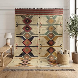 May include: A large rectangular area rug featuring a cream-colored base with a repeating geometric pattern in shades of red, navy blue, mustard yellow, green, and gray. The rug has a fringed edge at the top and bottom and a horizontal striped border.