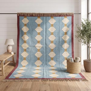 May include: A rectangular area rug featuring a light blue field with a repeating pattern of cream-colored diamonds connected by vertical mustard yellow bars. The rug is framed by a dark red border and finished with natural tan fringe on both ends.