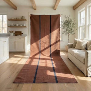 May include: A large, rectangular rug in a warm, reddish-brown hue with three vertical navy blue stripes. The rug is displayed indoors, with natural light illuminating its textured surface. The rug is made of a woven material.