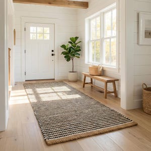 May include: An interior shot of a hallway with a patterned rug in black and cream. The rug lies on a wooden floor, leading to a white door. A wooden bench and a potted plant are also visible, creating a welcoming space.