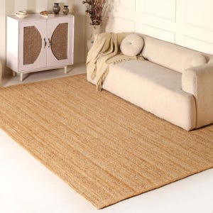 May include: A rectangular, natural-toned jute rug with a woven texture. The rug is placed on a white floor, with a cream-colored sofa and a white cabinet in the background. The rug adds a warm, earthy element to the room.