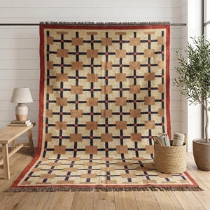 Può includere: Un tappeto rettangolare con sfondo color crema e un motivo a griglia ripetuto di linee blu navy scuro e quadrati color marrone chiaro. Il tappeto è incorniciato da un bordo rosso solido e rifinito con frange marrone scuro alle estremità.