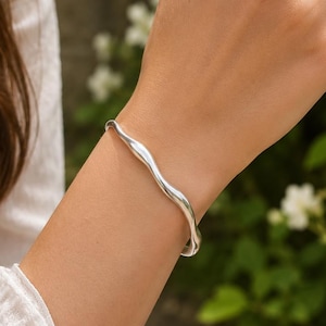 Pode incluir: Uma pulseira de prata polida com um design ondulado e suave no pulso, combinada com um anel de prata delicado com uma pequena cravação floral de várias pedras no dedo, ambos exibindo um acabamento metálico brilhante.
