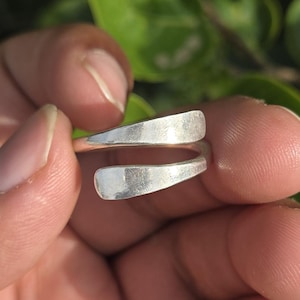 Può includere: Anello color argento dal design minimalista. L'anello presenta due fasce parallele e curve che non si collegano completamente, creando un aspetto aperto e moderno. L'anello è mostrato su uno sfondo di fogliame verde.