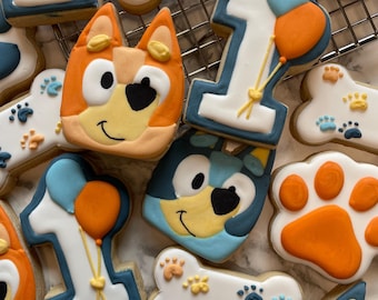Op de afbeelding: Een verzameling versierde koekjes met blauwe en oranje tekenfilmhondengezichten, het cijfer één met ballonnen, witte botvormen met kleine pootafdrukken en oranje pootkussentjes, gerangschikt op een metalen rooster.