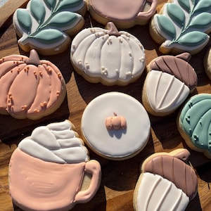 Könnte beinhalten: Eine Sammlung dekorierter Kekse, angeordnet auf einem Holztablett. Die Kekse zeigen herbstliche Designs, darunter Kürbisse, Eicheln, Blätter und Kaffeetassen, in Orange-, Weiß-, Braun- und Grüntönen.