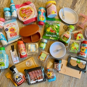 May include: A collection of miniature toy food items including bags of rice, crepes, and spring mix, various plastic bottles of sauces like BBQ and ketchup, small containers of sprinkles, chocolate chips, and snacks, plus tiny plates, forks, and cutting boards.