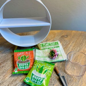 May include: A white circular shelf, two small packets labeled Sandy Mix and Rocky Mix by Happy Gro Co, a small green and pink succulent, a clear plastic cup, a miniature shovel, and a paper instruction sheet titled Mini Pots.
