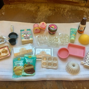 May include: A collection of miniature baking supplies, including a cake mix box, cookie dough packets, and various baking pans. There are also small containers of frosting and a bottle of syrup. The items are arranged on a white cloth.