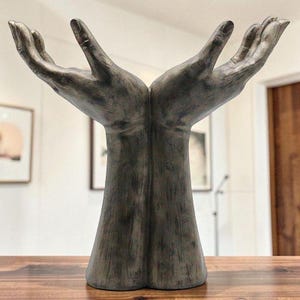 May include: A bronze-effect sculpture depicting two hands with open palms, reaching upwards. The sculpture sits on a wooden surface, with a blurred background of a room and framed pictures. The hands have detailed fingers and textured surfaces.
