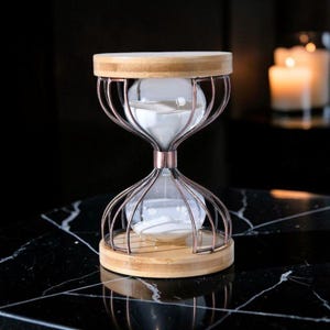 May include: An hourglass with a wooden top and base, and a copper-colored metal cage. The glass bulbs contain white sand. The hourglass is set on a black marble surface, with lit candles in the background.