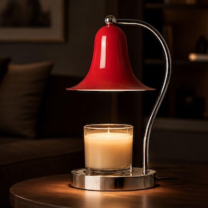 May include: A candle warmer lamp featuring a glossy red bell-shaped shade and a polished chrome curved arm. The base is circular and metallic, supporting a clear glass jar candle filled with white wax that is illuminated from above by the lamp.
