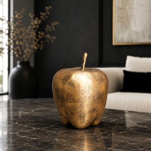 May include: A decorative apple sculpture featuring a metallic gold finish with dark, distressed, and textured detailing throughout the surface. The apple has a smooth, upright stem extending from the top center and a rounded, bulbous base.