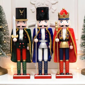 May include: Three wooden nutcracker figures standing in a row. The left figure wears a green cape and black hat with a sword. The centre figure wears a blue cape and black fur-style hat with a spear. The right figure wears a red cape and gold crown.