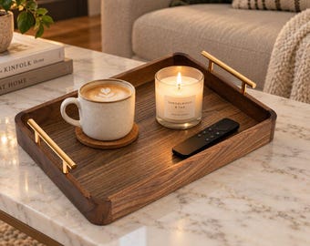 Könnte beinhalten: Ein rechteckiges Serviertablett aus dunklem Holz mit polierten goldfarbenen Metallgriffen. Auf dem Tablett stehen eine gesprenkelte Keramiktasse mit einem Latte, ein runder Korkuntersetzer, eine Glas-Duftkerze mit Aufschrift Sandalwood & Oak und eine Fernbedienung.