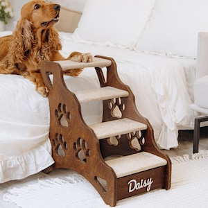 May include: A wooden pet step with three beige carpeted steps and paw print cutouts. The step is personalized with the name "Daisy" on the front. A brown Cocker Spaniel dog is resting on a white bed next to the step.