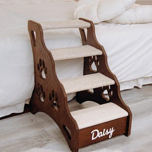 Puede incluir: Un escalón de madera para mascotas con tres escalones cubiertos de moqueta beige. El marco marrón oscuro presenta recortes de huellas de patas y el nombre "Daisy" en escritura blanca. Diseñado para ayudar a las mascotas.