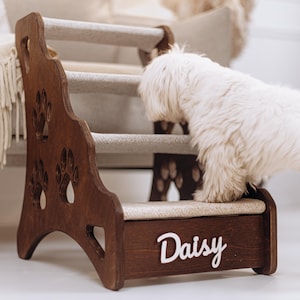 Puede incluir: Un escalón de madera para mascotas de color marrón oscuro con tres escalones cubiertos de moqueta beige. Los paneles laterales tienen recortes de huellas de patas. El nombre "Daisy" está en letras blancas en la parte delantera. Un perro blanco pequeño está usando los escalones.