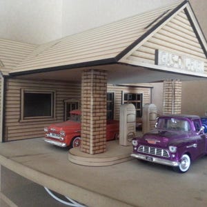 Puede incluir: Modelo en miniatura de una gasolinera vintage con dos camionetas cl&aacute;sicas. La gasolinera tiene un exterior de madera con el texto "GAS-OIL-FUEL" en el letrero. Una camioneta es roja, la otra es morada.