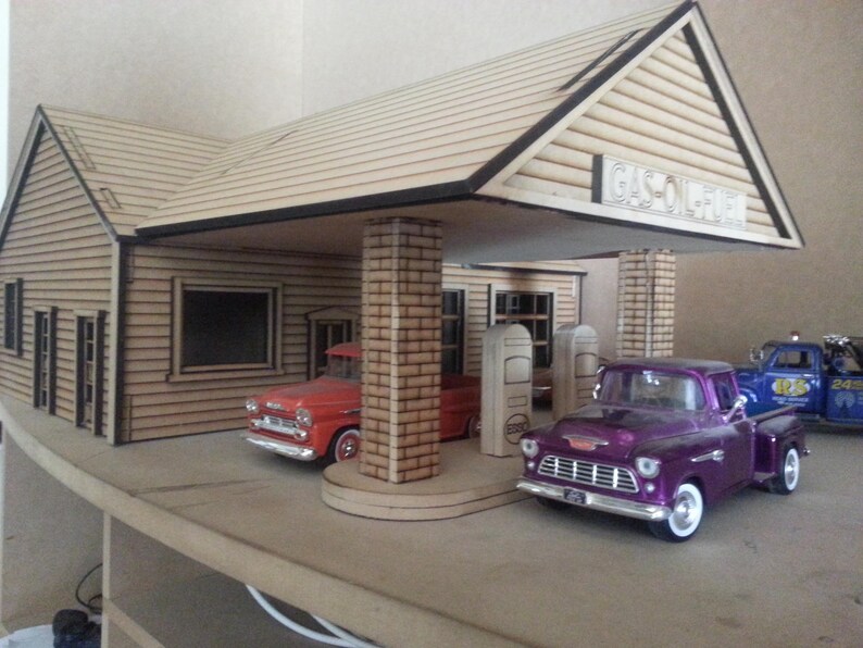 Puede incluir: Modelo en miniatura de una gasolinera vintage con dos camionetas cl&aacute;sicas. La gasolinera tiene un exterior de madera con el texto "GAS-OIL-FUEL" en el letrero. Una camioneta es roja, la otra es morada.