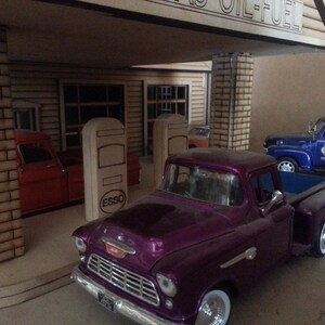 Puede incluir: Maqueta de gasolinera vintage con una camioneta morada en primer plano. La gasolinera tiene un letrero que dice "GAS OIL-FUEL". Otros veh&iacute;culos incluyen una camioneta roja y una gr&uacute;a azul.