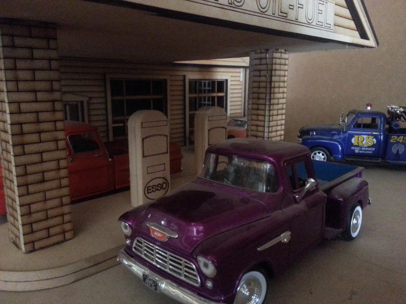 Puede incluir: Maqueta de gasolinera vintage con una camioneta morada en primer plano. La gasolinera tiene un letrero que dice "GAS OIL-FUEL". Otros veh&iacute;culos incluyen una camioneta roja y una gr&uacute;a azul.