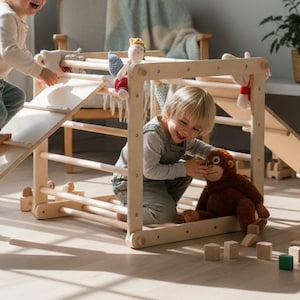 Op de afbeelding: Een houten klimframe met een helling en ladder, ontworpen voor kinderspel. Twee kinderen interageren met de structuur, de ene klimt de helling op en de andere speelt in het frame met een knuffel. Het frame is gemaakt van lichtgekleurd hout.