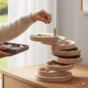 May include: A light wood jewellery organiser with multiple circular trays stacked in a spiral design. The organiser sits on a wooden dresser. A person is placing a necklace on the top tray. A small tray with jewellery is in the person's other hand.