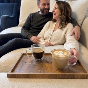 Op de afbeelding: Een rechthoekig houten dienblad met een donkerbruine afwerking met twee doorzichtige dubbelwandige glazen mokken. De ene mok bevat zwarte koffie, terwijl de andere een latte bevat met een hartvormig schuimpatroon op het oppervlak.