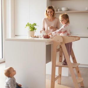 Puede incluir: Una torre de aprendizaje de madera natural con un niño de pie sobre ella, alcanzando un cuenco de fresas. Una mujer está cerca, sosteniendo una taza. Un bebé está en el suelo mirando hacia arriba. La cocina tiene una encimera y armarios blancos.