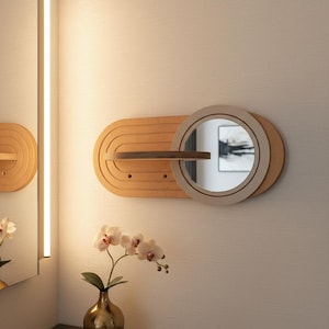 Op de afbeelding: Een wandspiegel van hout met een cirkelvormig glazen middenstuk in een lichtgekleurde, ovale houten achterplaat. Het ontwerp bevat een horizontale houten plank die over het midden loopt, met gegraveerde concentrische boogpatronen.