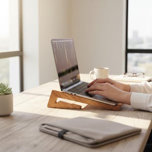May include: A light brown wooden laptop stand with a triangular side profile, supporting a silver laptop on a desk. The stand features a smooth, natural wood grain finish and an open design that elevates the laptop for an angled viewing position.